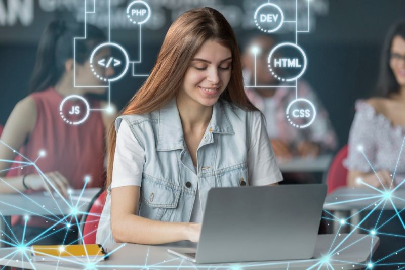 Young woman learning web development course on laptop with programming icons for HTML, CSS, PHP, JavaScript in a classroom setting